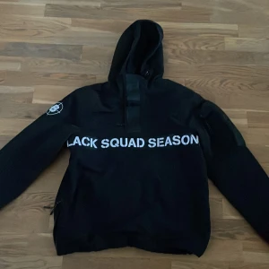 Svart jacka med text - Säljer en svart jacka med texten 'BLACK SQUAD SEASON' tryckt över bröstet. Jackan har en huva och dragkedja på sidan. Perfekt för en casual look.