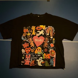 Santo t-shirt  - Cool svart t-shirt med ett färgglatt och lekfullt tryck på framsidan som inkluderar texten 'HOPE' och olika figurer. Baksidan har texten 'YOU'RE NOT SICK OF ME YET?' i olika färger. Perfekt för en unik och uttrycksfull stil.