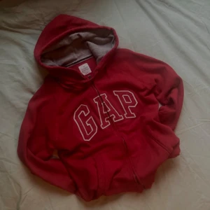 vintage Röd hoodie med  ”GAP” på - Säljer en jättesnygg röd hoodie  med dragkedja och stor logga på framsidan. Den har en mysig grå insida i huvan och är perfekt för kyliga dagar. Passar både till vardags och avslappnade tillfällen.