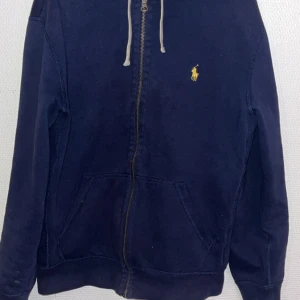 Mörkblå huvtröja från Ralph Lauren - Säljer en snygg mörkblå huvtröja från Ralph Lauren med dragkedja och klassisk logga på bröstet. Tröjan har långa ärmar och en praktisk huva med snören. Perfekt för en avslappnad stil.