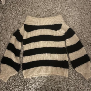 Randig stickad tröja - Säljer en mysig stickad tröja med breda svarta och beige ränder. Tröjan har en offshoulder-design och långa ärmar med ribbade muddar. Perfekt för kyliga dagar! 😍😍