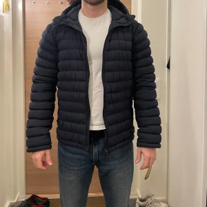 Svart pufferjacka från J.Lindeberg - Säljer en stilren mörkblå pufferjacka från J.Lindeberg. Jackan har en dragkedja framtill och en praktisk huva. Perfekt för kyliga dagar med sin quiltade design som håller värmen. Passar bra till både jeans och chinos.