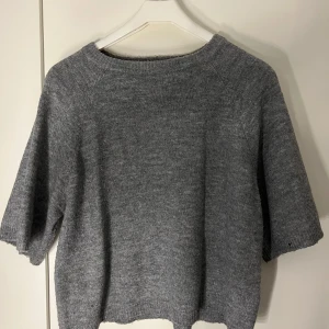 Grå stickad tröja - Mysig grå stickad tröja med korta ärmar. Perfekt för lager-på-lager under kyligare dagar. Enkel och stilren design som passar till det mesta.