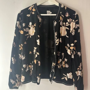 Tjocktröja Saint Tropez  - Svart tjocktröja med blommönster. Tjocktröjan har en dragkedja framtill och ribbade muddar vid ärmslut och nederkant. Från Saint Tropez. Storlek M