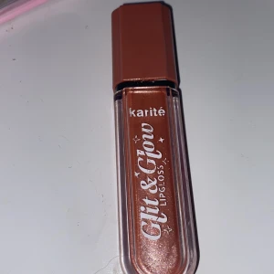 Karité Glit & Glow Lipgloss - Snyggt glittrande läppglans från Karité i en varm, kopparfärgad nyans. Perfekt för att ge läpparna en skimrande och fyllig look. Kommer i en praktisk tub med applikator för enkel applicering.