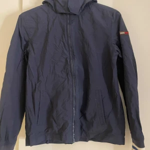 Blå vindjacka från Tommy Hilfiger - Snygg blå vindjacka från Tommy Hilfiger med dragkedja och huva. Jackan har en diskret logga på ärmen och är perfekt för blåsiga dagar. Den är långärmad och har en klassisk passform.