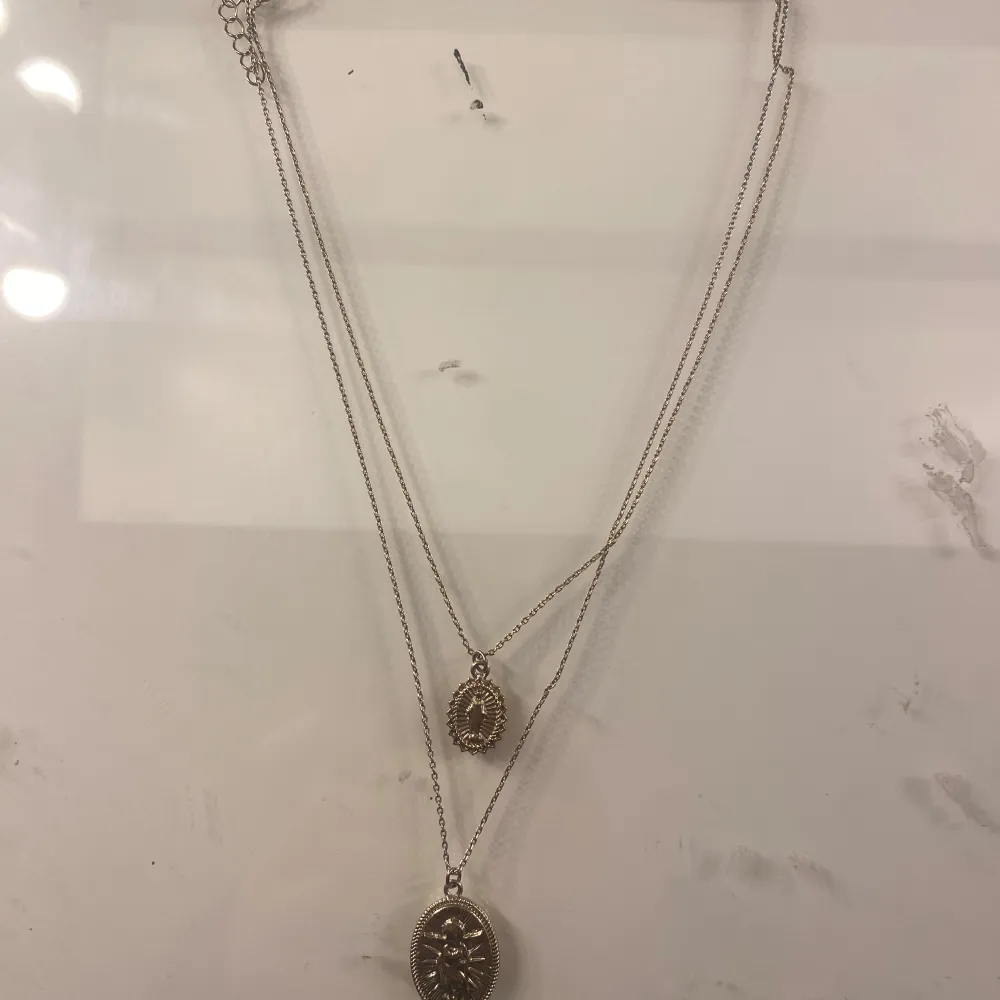 Elegant dubbelhalsband i guld med två hängsmycken. Det ena hängsmycket har en oval form med en stjärndesign, medan det andra är mindre och rundat. Perfekt för att lägga till en touch av stil till din outfit.. Asusteet.
