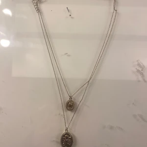 Dubbelhalsband i guld - Elegant dubbelhalsband i guld med två hängsmycken. Det ena hängsmycket har en oval form med en stjärndesign, medan det andra är mindre och rundat. Perfekt för att lägga till en touch av stil till din outfit.