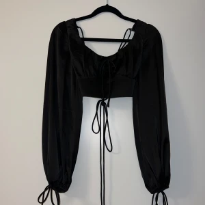 Svart långärmad topp från Kiss The Sky - Snygg svart topp från Kiss The Sky i storlek XS. Toppen har långa ärmar och en trendig snörning framtill. Perfekt för en kväll ute eller en speciell tillställning. Andra bilden är på ryggen, så snyggt. Aldrig använd!