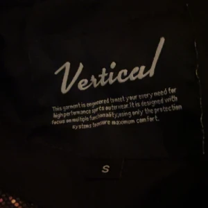 Svart jacka från Vertical - Säljer en svart jacka från Vertical, perfekt för sportiga aktiviteter. Jackan har en huva och dragkedja framtill. Insidan är designad för maximal komfort och funktionalitet.