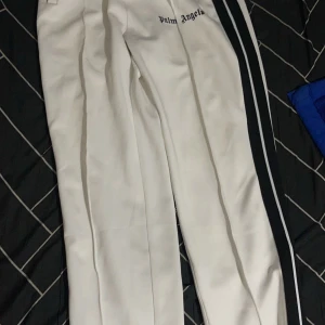  Palm Angels byxor - Snygga vita byxor från Palm Angels med svarta ränder längs sidorna. Byxorna har en modern design med dragkedjor vid bensluten och en bekväm passform. Perfekta för en stilren look. 