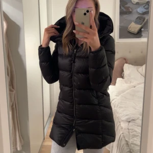 Svart dunjacka från Parajumper - Säljer en svart dunjacka från Parajumper med huva och dragkedja. Jackan är lång och perfekt för kalla dagar. Den har en stilren design med märkets logga på ärmen.