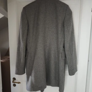 Grå kappa med knappar - Elegant grå kappa med dubbelknäppning och stilrena knappar. Långärmad design med klassisk passform, perfekt för kyligare dagar. Tidlös och sofistikerad stil.