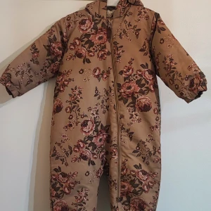 Blommig overall med huva - Detta är en storlek för barn86cm passar till 18-24 månader Säljer en söt beige overall med blommigt mönster i rosa och grönt. Overallen har en praktisk dragkedja framtill och en mysig huva. Perfekt för kyligare dagar.