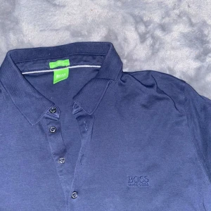 Mörkblå pikétröja från Hugo Boss - Snygg mörkblå pikétröja från Hugo Boss med klassisk krage och knappar framtill. Perfekt för en stilren look. Märkeslogga diskret broderad på bröstet. Storlek Xxl, passform är dock L. 