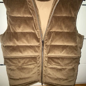 Beige manchester dunväst från Stenströms - Någon intresserad att köpa min Snygg beige manchester dunväst från Stenströms med dragkedja och huva. Perfekt för höst och vinter. Västen har en klassisk look med praktiska fickor framtill. 