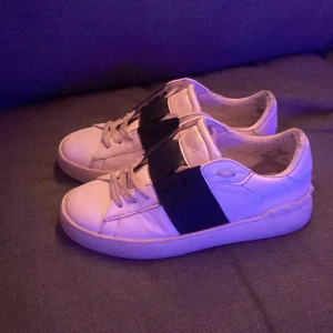 Valentino open skor - Snygga Valentino Garavani sneakers i vitt med en bred svart rem över snörningen. Skorna har en klassisk design med rund tå och är tillverkade i skinn. Perfekta för en stilren look. Gör jätte gärna byten