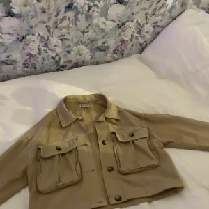 Beige croppad jacka med fickor - Snygg beige croppad jacka med stora fickor och knappar framtill. Perfekt för en avslappnad stil. Jackan har en klassisk krage och är tillverkad i ett lätt material.