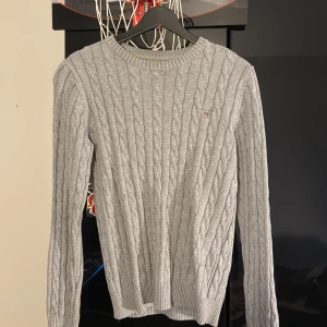 Grå stickad tröja från GANT - Säljer en stilren grå stickad tröja från GANT i premium bomull. Tröjan har ett klassiskt flätmönster och rund halsringning. Perfekt för kyligare dagar.