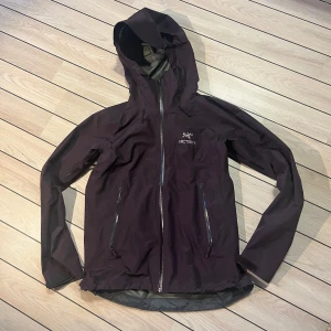 Mörk lila vattentät skaljacka från Arc'teryx - Säljer en stilren mörklila Arc'teryx jacka med huva och vattentäta dragkedjor. Jackan är goretex har en minimalistisk design med logotyp på bröstet och är perfekt för utomhusaktiviteter. Den är tillverkad i ett slitstarkt material som skyddar mot väder och vind. Den är vattentät och ventilerande, 2-vägs dragkedja, justerbar huva med kort skärm, 2 handfickor m dragkedja, kardborrejustering i ärmslut, hög krage. Mycket hög kvalitet. Använd några gånger. Nypris 4799kr