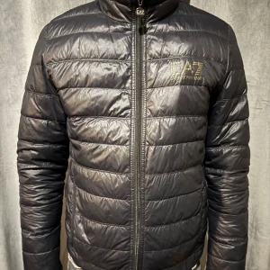 Svart pufferjacka från EA7 - Snygg svart pufferjacka från EA7 med dragkedja framtill. Jackan är lite solblekt samt guldloggan. Men har inga skador eller dylikt så den har mycket kvar att ge.