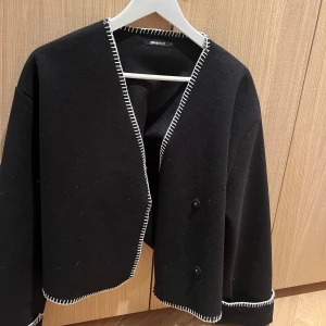 Svart kavaj/jacka från Ginatricot - Snygg svart kavaj från Ginatricoy med vita kontrastsömmar. Kavajen har en croppad passform och knappar framtill. Perfekt för en stilren look, den är lite tunnare.