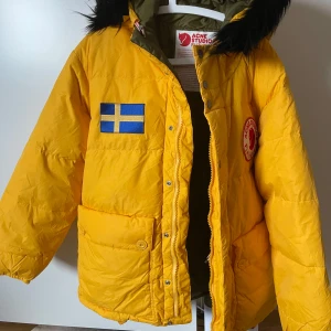Gul dunjacka från Acne Studios x Fjällräven - Säljer en cool gul dunjacka från Acne Studios x Fjällräven med svensk flagga och logotypdetaljer. Jackan har en avtagbar huva med svart päls och knäppning framtill. Perfekt för kalla dagar!