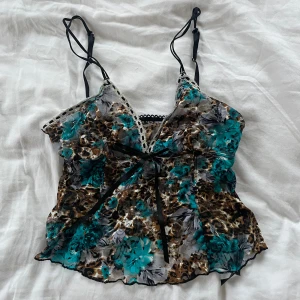 Blommigt linne med spetsdetaljer - Säljer ett blommigt linne med tunna axelband och spetsdetaljer. Linnet har en knytning framtill och ett färgglatt mönster i turkos, brun och svart.  Endast provat, därför i nyskick!