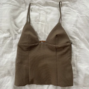 Brunt linne med smala axelband - Snygg brunt linne med smala axelband och en liten spänndetalj framtill. Toppen har en figurnära passform och ett diskret rutmönster. Perfekt för en stilren look.