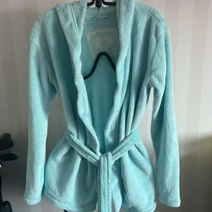 Ljusblå morgonrock från H&M - Mysig ljusblå morgonrock från H&M i mjukt material. Den har en huva och knyts med ett bälte i midjan. Perfekt för att hålla sig varm och bekväm hemma.