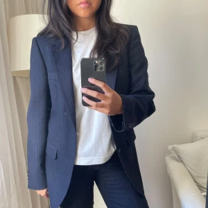 Mörkblå kavaj med diskreta ränder - Mörkblå kostym med diskreta ränder. Perfekt för både vardag och fest! Kavajen har en klassisk passform med långa ärmar och knappar framtill. Den är i bra skick och passar till många olika outfits.