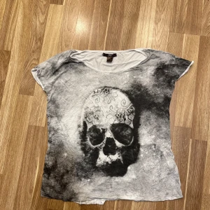 T-shirt med dödskallemotiv - T-shirt från Ellos med ett stort dödskallemotiv på framsidan. T-shirten är i en grå och svart färgskala med en unik, lite sliten look. Perfekt för en avslappnad stil eller för att ge lite edge till din outfit. Storlek 48/50