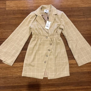 Beige rutig kavajklänning från NA-KD - Säljer en superfin beige kavajklänning från NA-KD med ett stilrent rutmönster. Klänningen har långa ärmar och knappar framtill, vilket ger en elegant look. Perfekt för både vardag och fest!