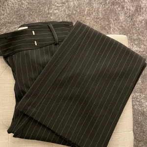 Svarta randiga kostymbyxor från Clockhouse - Säljer ett par stiliga svarta kostymbyxor från Clockhouse med tunna vita ränder. Byxorna har en klassisk design med bälteshällor och en diskret ficka bak. Perfekta för både formella tillfällen och kontoret. Storleken är inte angiven, men passar M