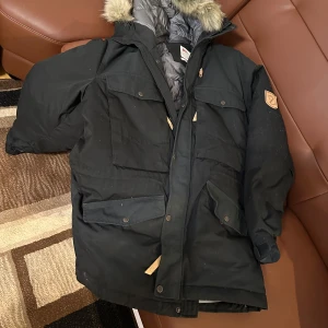 Svart parkajacka från Fjällräven - Säljer en svart parkajacka från Fjällräven i G-1000 material. Jackan har en avtagbar huva med pälsimitation och flera praktiska fickor med knappar. Perfekt för kalla dagar med sin långa design och varma foder. Helt nytt 