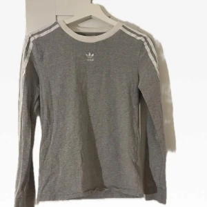 Grå långärmad tröja från Adidas - Säljer en stilren grå långärmad tröja från Adidas med klassiska vita ränder längs ärmarna. Perfekt för en sportig look eller avslappnad stil. Adidas-loggan pryder bröstet. Passar till många tillfällen!
