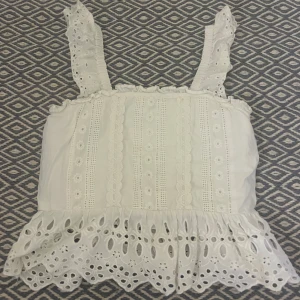 Vit spetsprydd topp - Fin vit topp med spetsdetaljer och volangkant. Axelbanden är dekorerade med spets och toppen har en elastisk rygg för bättre passform. Perfekt inför sommaren!
