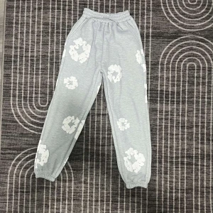 Grå hoodie och byxor med blommönster - Säljer ett set med en grå hoodie och matchande byxor, båda med ett vitt blommönster. Hoodien har en stor ficka framtill och byxorna har resår i midjan för extra komfort. Perfekt för en avslappnad stil.