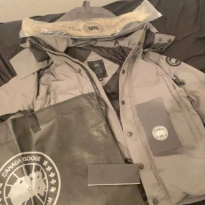 Grå dunjacka från Canada Goose - Säljer en stilren grå dunjacka från Canada Goose. Jackan har en hög krage och flera praktiska fickor. Perfekt för kalla dagar med sin varma och skyddande design. Den har en dragkedja och knappar för extra skydd mot väder och vind.