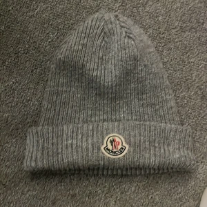 Grå ribbad mössa från Moncler - Snygg grå ribbad mössa från Moncler med uppvikt kant och broderad logotyp framtill. Perfekt för kyliga dagar och ger en stilren look.