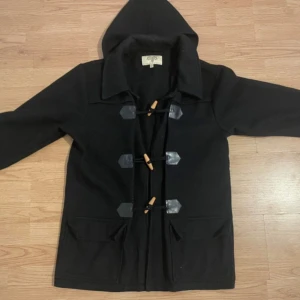 Svart duffeljacka från Euroa Clothing Co. - Säljer en svart duffeljacka från Euroa Clothing Co. med huva och klassiska träknappar. Jackan har långa ärmar och två stora fickor framtill. Perfekt för kyliga dagar.