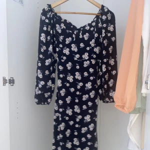 Blommig klänning med långa ärmar - Säljer en söt blommig klänning med svart bas och vita blommor. Klänningen har långa ärmar och en smickrande passform med smockdetaljer vid bysten. Perfekt för en somrig look!