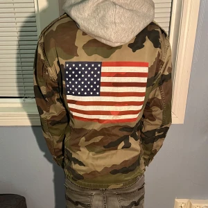 Camo overshirt - Snygg camouflagejacka med en stor amerikansk flagga på ryggen. Jackan har dragkedja och flera praktiska fickor. Perfekt för en cool och avslappnad stil.