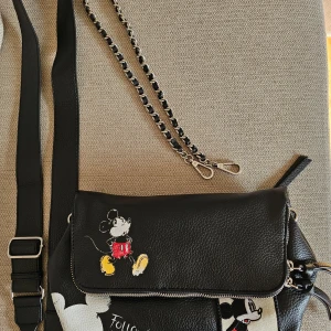 Svart axelväska med Musse Pigg från Desigual - Upper flap with magnetic closure An outer pocket and a small pocket inside, both with zipper Inner compartment divided into three spaces, one of them with zipper Mickey Mouse prints with worn effect Patch of a Mickey Mouse comic panel Big pullers in leather effect fabric The flap is a big pocket with its own zip fastener Includes two interchangeable straps: one in leather effect fabric and the other one is a chain Measurements: 25.5 (l.), 12.6 (w.), 13.5 (h.) cm.