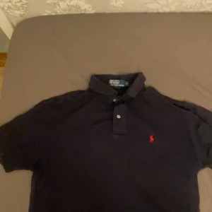 Svart pikétröja från Ralph Lauren - Säljer en klassisk svart pikétröja från Ralph Lauren med den ikoniska röda loggan på bröstet. Tröjan har korta ärmar och en stilren krage med knappar. Perfekt för en avslappnad men stilfull look.