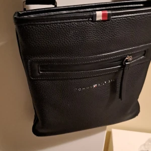 Svart axelväska från Tommy Hilfiger - Snygg svart axelväska från Tommy Hilfiger i skinn med dragkedja framtill. Väskan har en justerbar axelrem och en stilren design med märkets logotyp på framsidan. Perfekt för att bära dina dagliga nödvändigheter med stil.