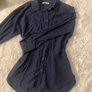 Mörkblå skjorta med knappar - Snygg mörkblå skjorta med en krinklad textur och knappar framtill. Skjortan har långa ärmar och en klassisk krage, perfekt för en stilren look.