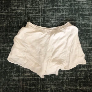 Vita shorts med spetsdetalj - Säljer ett par vita shorts med spetsdetalj i midjan. De är löst sittande och perfekta för varma dagar. Materialet ser mjukt och bekvämt ut, vilket gör dem till ett skönt val för avslappnade tillfällen.