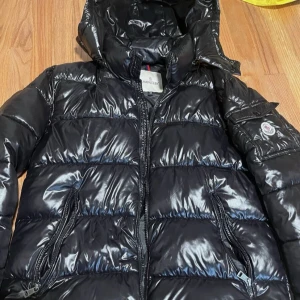 Svart pufferjacka från Moncler - Säljer en snygg svart pufferjacka från Moncler med glansig yta och huva. Jackan har dragkedja och två fickor framtill. Perfekt för kyliga dagar.