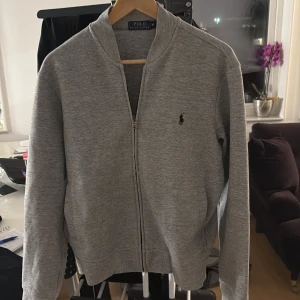 Grå Ralph Lauren zip tjocktröja  - Säljer en stilren grå hoodie från Ralph Lauren med dragkedja och det klassiska logotypbroderiet på bröstet. Perfekt för en avslappnad look. Den har långa ärmar och är tillverkad i ett mjukt material. Den är oanvänd och jag fick den i present men passade tyvärr inte mig 